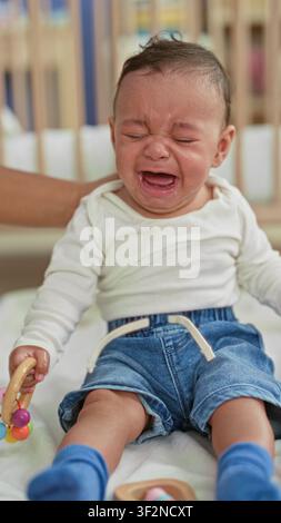 Bambino ispanico che piange, tiene un sonaglio di legno mentre mani adulte asciugano le lacrime in una culla di edificio; cure di consolazione in caso di distress. Foto Stock