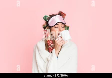Donna assonnata in arricciacapelli e maschera per il sonno avvolto in coperta e bevande caffè da tazza Foto Stock