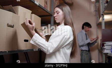 Giovane donna che etichetta scatole d’archivio in un magazzino con un collega sullo sfondo Foto Stock
