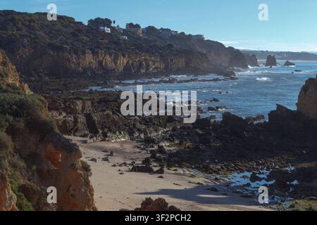 Le onde si infrangono contro scogliere rocciose lungo la costa. Il litorale presenta un'area sabbiosa, mentre il sole splende sulla scena. Alcune case sono visibili Foto Stock
