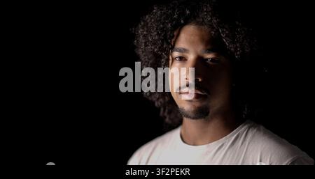 Giovane uomo con capelli ricci e barba, illuminato da un faretto a luce bassa su uno sfondo scuro Foto Stock