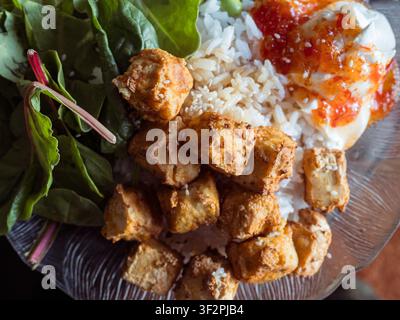 Un macroscatto dall'alto di una ciotola di cibo nutriente illuminata dal sole, ripiena di cubetti di tofu croccanti, riso bianco, verdure a foglia e salsa. Foto Stock