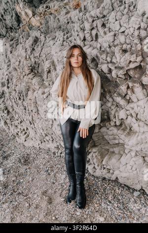 Ritratto di moda su scogliera di una giovane donna appoggiata a un muro di roccia ruvida su una spiaggia di ciottoli Foto Stock