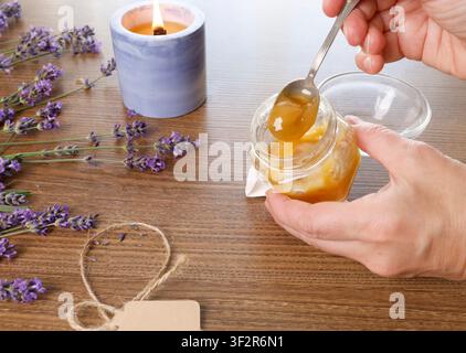Creatore di contenuti creativi che raccoglie miele di lavanda crudo biologico da un piccolo barattolo di vetro su una scrivania di legno rustica vicino a una candela profumata accesa per l’olismo consapevole Foto Stock