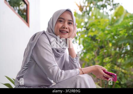 Allegra donna musulmana asiatica con hijab e apparecchio dentale, che tiene uno smartphone mentre si rilassa all'aperto Foto Stock