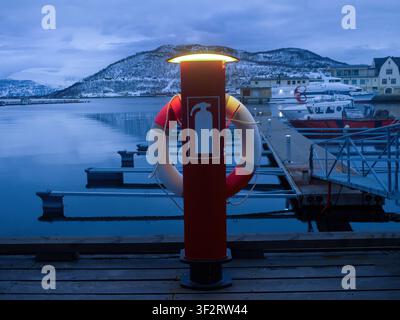 Una foto atmosferica di un palo di sicurezza rosso sul molo, con un anello di salvataggio e un simbolo di estintore illuminato da una luce calda incorporata. Il primo piano Foto Stock