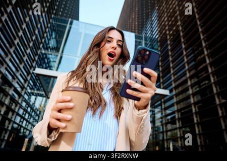 Giovane donna che sembra scioccata e stupita mentre controlla i contenuti sul suo telefono cellulare, tenendo una tazza di caffè in città Foto Stock