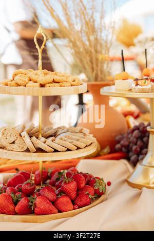 Un primo piano a fuoco selettivo di un tavolo da catering splendidamente decorato con fragole fresche e mature, pane a fette e biscotti su un vassoio di legno a più livelli Foto Stock
