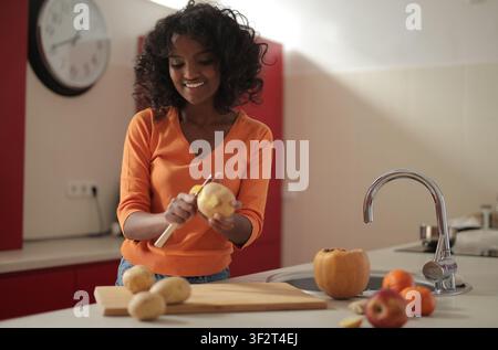 Giovane donna nera felice che sbuccia le patate in cucina Foto Stock