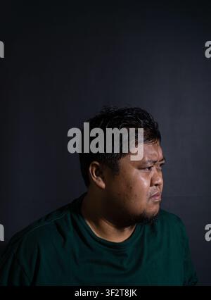 Ritratto di profilo ravvicinato di un uomo indonesiano adulto serio con una camicia verde scuro che guarda lontano su uno sfondo nero intenso. Il soggetto dis Foto Stock
