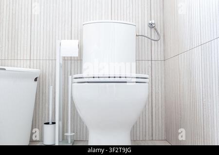 Vista frontale di una moderna vasca da bagno in ceramica bianca con coperchio chiuso, portarotolo, spazzolino e piccolo cestino della spazzatura su pareti piastrellate chiare in un Foto Stock