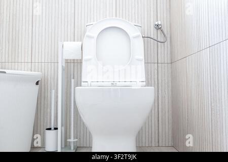 Vista frontale di una moderna tazza di toilette in ceramica bianca con seduta e coperchio aperti, portarotolo, spazzolino e piccolo cestino della spazzatura su uno sfondo di parete piastrellata luce Foto Stock