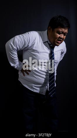 Uomo asiatico adulto affaticato che tiene la parte bassa della schiena dolorante, si appoggia all'indietro con un'espressione facciale smorfiosa su uno sfondo nero solido. L'ov Foto Stock