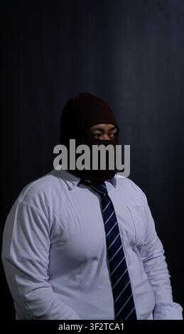 Uomo adulto dell'Asia sudorientale che indossa una maschera balaclava in maglia marrone, in piedi su uno sfondo nero scuro. La figura misteriosa è vestita in modo formale Foto Stock