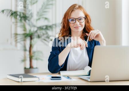 Una donna d'affari dai capelli rossi sorride mentre lavora su un computer portatile alla scrivania in un ufficio luminoso Foto Stock