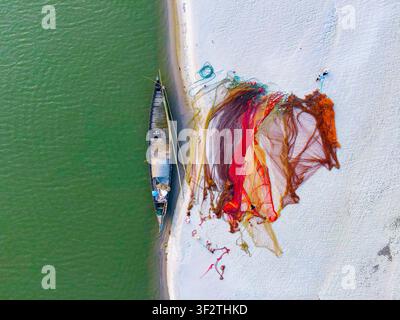 Vista aerea di una tradizionale barca da pesca in legno ormeggiata su una riva sabbiosa con reti da pesca vivaci arancioni e rosse stese ad asciugare a Kushtia, Kh Foto Stock