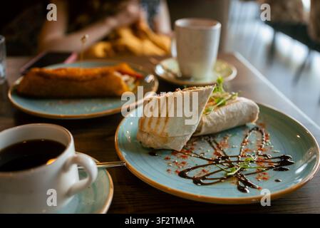 Involto di falafel vegano servito su un piatto con glassa al balsamico e semi di sesamo, che completa un pranzo vegetale in un caffè con caffè e un altro panino Foto Stock