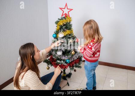 Famiglia che decora un piccolo albero di Natale, aggiungendo ornamenti colorati e fili luccicanti per il periodo delle festività Foto Stock