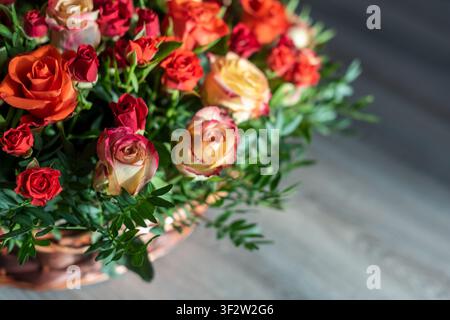Vista ravvicinata di un cesto di vimini pieno di rose colorate su uno sfondo grigio scuro. Rose rosso chiaro, rosso scuro, arancione e rosso-giallo con fogliame verde Foto Stock