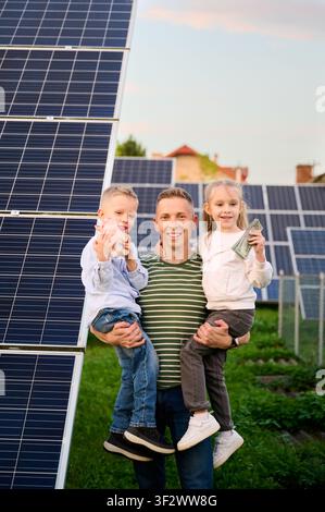 Padre che tiene due bambini vicino ai pannelli solari, i bambini mostrano un salvadanaio e denaro. Famiglia felice che investe nelle energie rinnovabili, concetto di risparmio, stile di vita sostenibile Foto Stock