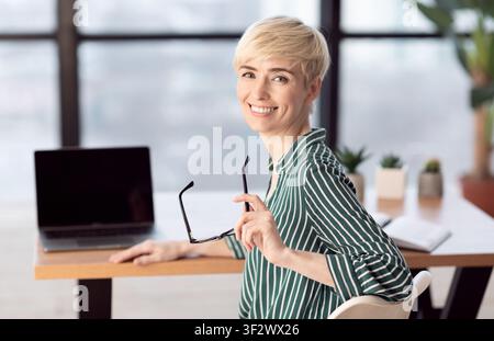 Felice Businesswoman Di Mezza Età Seduto Alla Scrivania In Ufficio Moderno Foto Stock