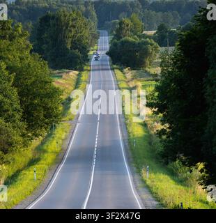 Automobili che percorrono una lunga strada rurale ondulata attraverso la campagna vicino a Viesīte, Lettonia. Foto Stock