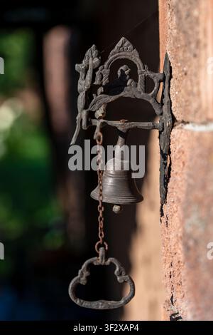 Campanello vintage in ghisa con catena su muro di pietra, primo piano. Campanello di porta in bronzo antico appeso a un muro di mattoni rustico con una staffa metallica decorativa a Foto Stock