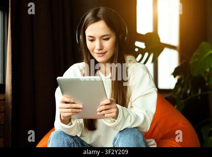 La ragazza moderna con tablet e cuffie funziona nella sedia a sacco Foto Stock