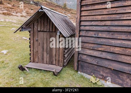 Piccola capanna di tronchi con tetto spiovente, situata su terreno erboso, adiacente a un muro più grande di una tradizionale baita in legno Foto Stock