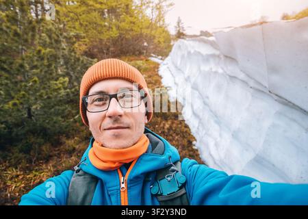 Giovane uomo che sorride alla telecamera, indossa un berretto e una giacca, in piedi vicino a un cumulo di neve durante un'escursione in montagna Foto Stock