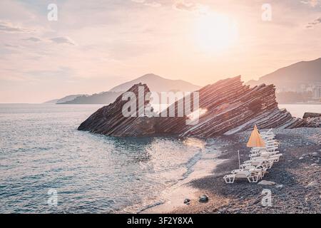 Il sole che tramonta sull'Adriatico, illuminando una formazione rocciosa stratificata unica e sdraio vuoti su una spiaggia di ciottoli in Montenegro Foto Stock