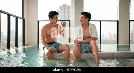 Coppia gay che si rilassa in piscina sul tetto della città, godendo di conversazioni e bevande Foto Stock