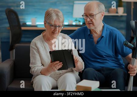 Coppia anziana guarda uno schermo del tablet nel soggiorno, godendosi il tempo libero insieme con la navigazione digitale e il relax domestico durante una tranquilla routine casalinga. Foto Stock