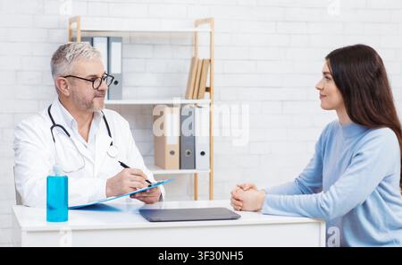 Uomo adulto in camice bianco e bicchieri fa note, parla con la paziente femminile al tavolo con disinfettante e laptop Foto Stock