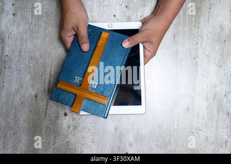 Vista dall'alto di mani che tengono una Bibbia blu e una croce di legno sopra un tablet bianco su uno sfondo grigio, rappresentando l'intersezione della tradizione Foto Stock