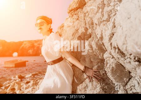 Donna vintage, signora costiera in abito bianco appoggiata sulle rocce durante il tramonto, catturando una nostalgia estiva onirica e vibrazioni vacanziere Foto Stock