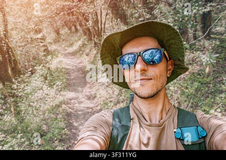 Giovane uomo con occhiali da sole e cappello che scatta un selfie mentre esplora un sentiero escursionistico in una foresta lussureggiante in una giornata soleggiata Foto Stock
