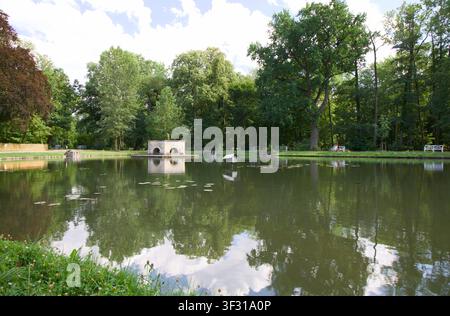 Stagno scenografico circondato da alberi verdi rigogliosi a Laxenburg, Austria, con una piccola struttura in pietra con aperture ad arco riflesse nell'acqua calma Foto Stock