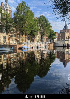 Vista estiva del canale Geldersekade ad Amsterdam con la torre storica Schreierstoren, alberi verdi e barche riflesse nell'acqua ferma Foto Stock