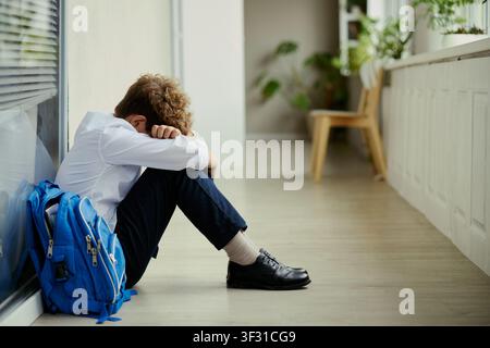 Ragazzino seduto per terra con la testa sulle braccia accanto allo zaino, che mostra tristezza, stress, bullismo o ansia scolastica. Utile per campagne di educazione, salute mentale dei bambini o consulenza Foto Stock