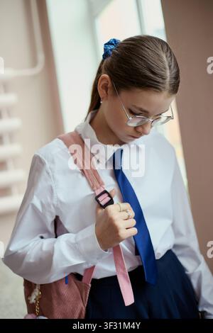Fotografia verticale di una scolara caucasica con occhiali, che tiene la cinghia dello zaino e guarda in basso mentre si prepara per le lezioni. Adatta per l'educazione, la routine scolastica, le campagne di apprendimento giovanile Foto Stock