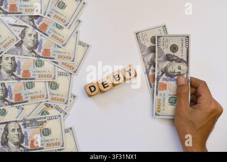 Una mano che tiene banconote da dollari USA accanto a blocchi di legno che formano la parola debito su sfondo bianco. Foto Stock