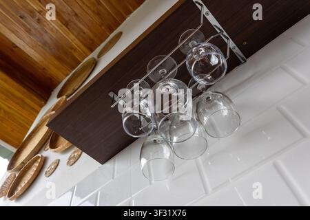 Un chiaro scaffale per bicchieri da vino sospeso sotto un armadio di legno con luce riflessa, dettaglio accogliente della cucina e piatti tessuti decorativi che aggiungono una texture rustica e w Foto Stock