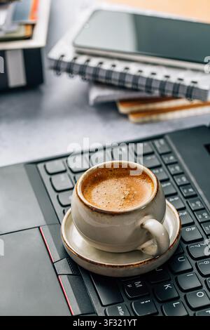 Una tazza di espresso è appoggiata su una tastiera del laptop accanto a quaderni e un telefono Foto Stock