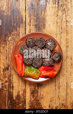 Fette di morcilla grigliate con peperoni rossi e verdi arrostiti, servite su un piatto di terracotta sopra un tavolo di legno stagionato. Foto Stock