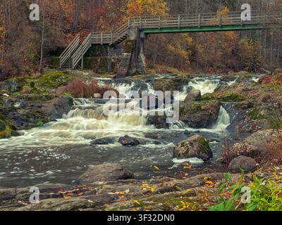 Kuusimäki, Finlandia - 20.10.2018: Acqua setosa scorre su pietre scure sotto un ponticello di legno stagionato in mezzo a una vegetazione autunnale vibrante Foto Stock
