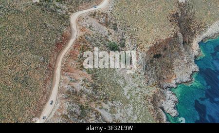Vista aerea di auto che percorrono una strada sterrata accidentata su una scogliera ripida verso la spiaggia di Balos sull'isola di Creta in Grecia durante una giornata estiva soleggiata in un paesaggio costiero selvaggio e scenografico Foto Stock