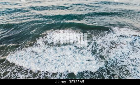 Ripresa aerea dall'alto di acqua oceanica di un blu profondo con un'onda schiumosa bianca che si diffonde sulla superficie del mare creando uno sfondo naturale texture durante una giornata estiva soleggiata Foto Stock