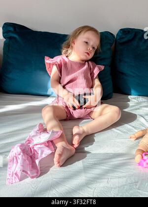 Bambino seduto su un letto che tiene una macchina giocattolo con bambole e vestiti alla luce naturale del sole. Le scelte dell'infanzia incoraggiano l'immaginazione, l'indipendenza e Foto Stock