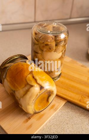 Funghi marinati fatti in casa in barattoli di vetro su tagliere di legno, conserva di funghi di bosco biologici, ambientazione rustica in cucina in primo piano Foto Stock